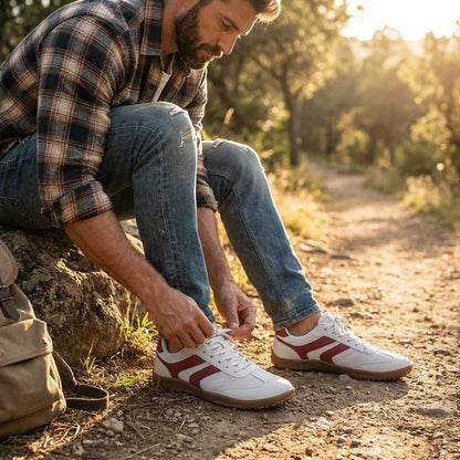 Man tying his white barefoot sneakers with red accents outdoors on a dirt path.